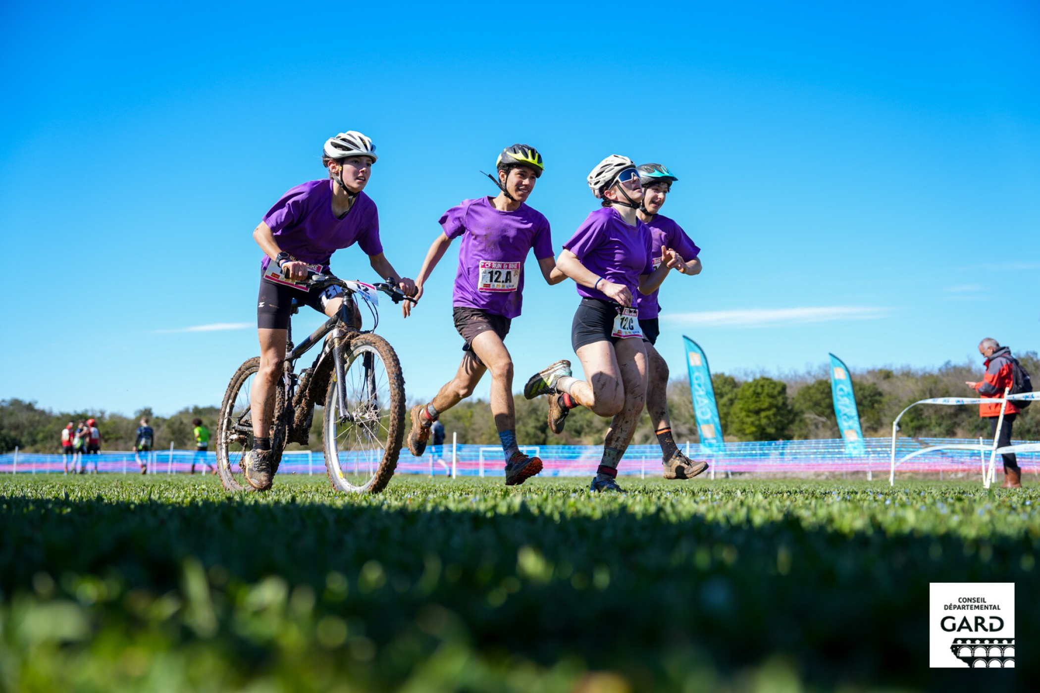 Photo de 3 jeunes athlètes, 1 en VTT, les 2 autres en course à pied sur une pelouse bien verte et un ciel bleu azur